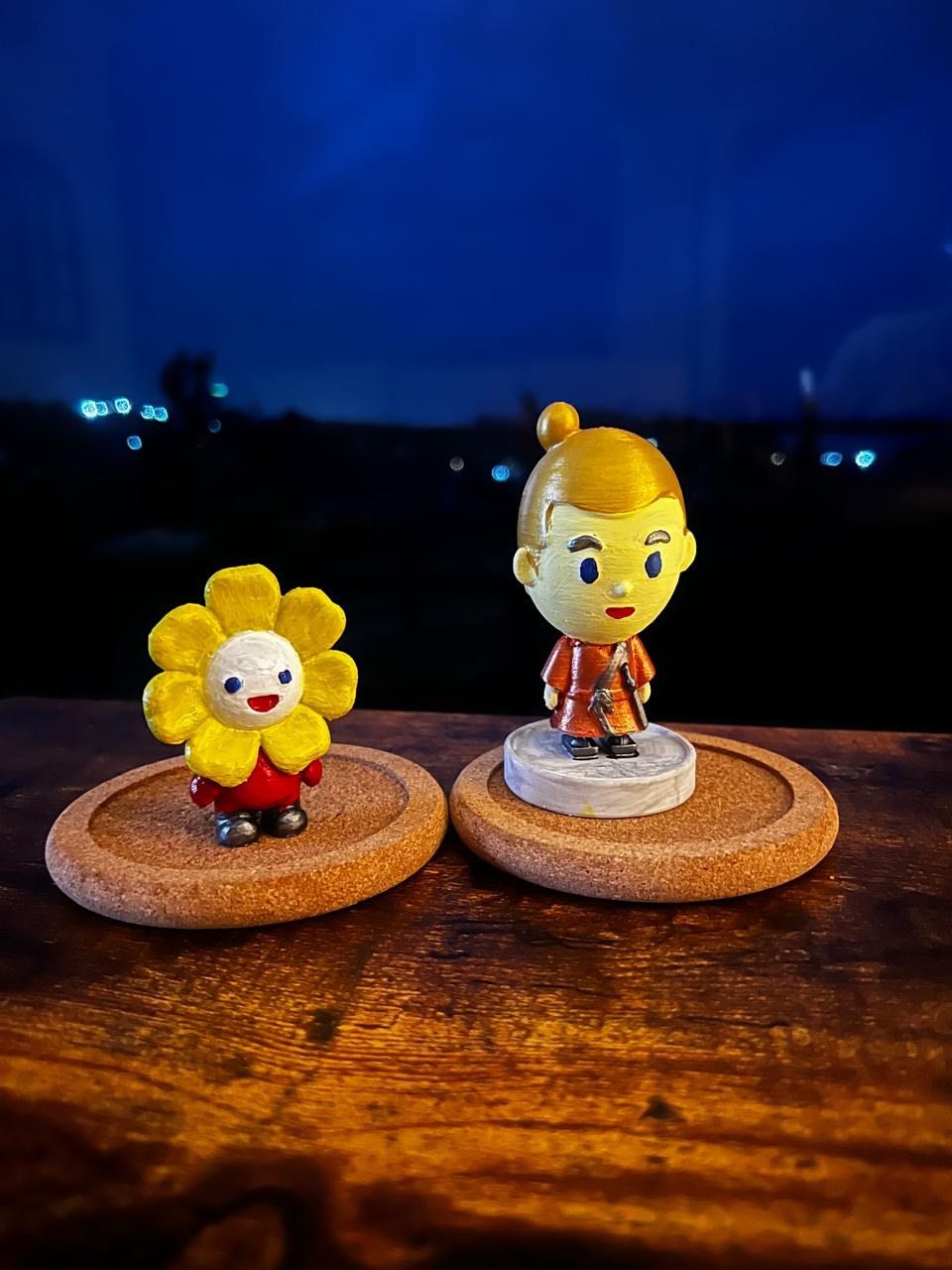 A flower Pop and a Pop with a top knot, painted, on cork coasters at dusk